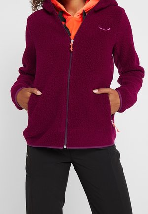 Persoon draagt een paarse fleecejas met ritssluiting en zakken en een oranje capuchontrui eronder, staand met handen in de zakken.