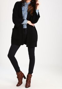 Svart ribbad cardigan med vida ärmar och framfickor, bärs över en denimskjorta och kombineras med mörka leggings och bruna ankelboots.