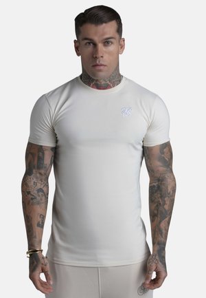 Man met tatoeages op armen en nek, gekleed in een strakke beige korte mouwen t-shirt met een klein wit logo op de borst, staand tegen een witte achtergrond.