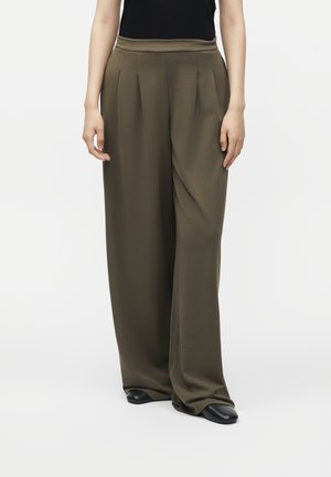 Personne portant un pantalon large plissé vert olive, des chaussures noires et un haut noir sans manches, debout devant un fond blanc uni.