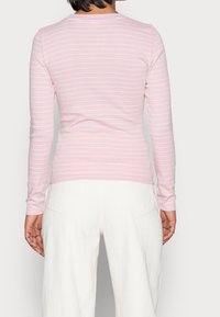 Top a maniche lunghe rosa a righe realizzato in tessuto morbido, caratterizzato da sottili righe bianche. Indossato con pantaloni bianchi a vita alta, che mostrano una silhouette aderente.