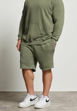 Mand iført olivengrøn sweatshirt og matchende shorts med hvide Nike-sneakers på grå gulv og ensfarvet baggrund.
