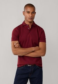 Bordová polo tričko s golierom, s trojzubkovým zapínaním a krátkymi rukávmi, vyrobené z hladkej látky. Nosené s tmavomodrými džínsami.