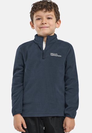 Jeune garçon portant une veste en polaire bleu marine avec fermeture éclair quart, arborant le logo "Jack Wolfskin" sur la poitrine, se tenant devant un fond uni.