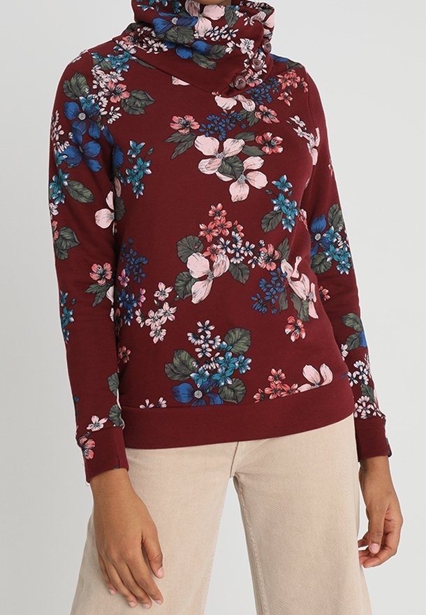Sweat-shirt à col roulé fleuri avec des fleurs roses, bleues et vertes sur un tissu bordeaux, porté avec un pantalon beige.