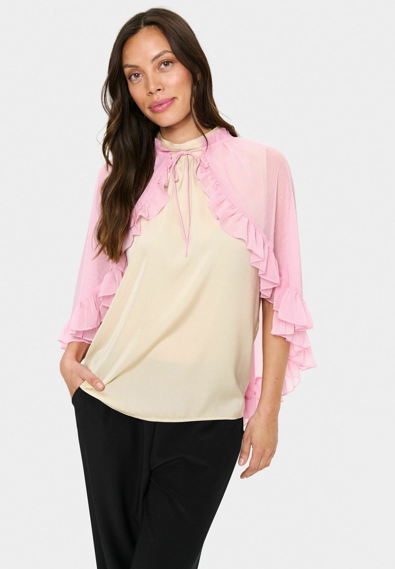 Lightweight, ruffled pink cape overlay with a tie closure, worn over a beige top and paired with black trousers. Smooth texture, flowing design.