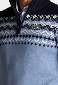 Gebreide pullover in gradient blauw met een donkerblauwe kraag. Bevat geometrische patronen in wit en marineblauw, een ritssluiting en een logo patch.