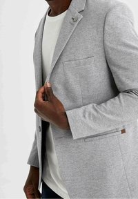 Blazer gris con una tela texturizada, de un solo botón, con bolsillo en el pecho y detalles en cuero. Llevado sobre una camisa de color claro.