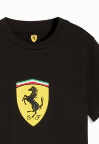 Černé tričko s kulatým výstřihem a žlutým logem Ferrari ve tvaru štítu, zobrazujícím skákajícího koně a zelené, bílé a červené pruhy nahoře.