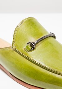 Mocassin en cuir vert avec un bout arrondi, présentant un accent en métal argenté sur le dessus et des détails de couture assortis.