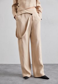 Beige weit geschnittene Hose mit glatter Textur, die ein einzigartiges Wickeldetail an der Taille und einen einzelnen schwarzen Knopf aufweist, kombiniert mit spitzen schwarzen Schuhen.
