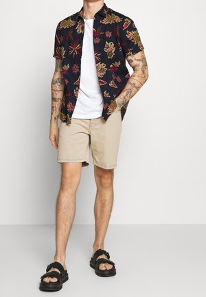 Homme aux bras tatoués portant une chemise noire à manches courtes à motifs floraux par-dessus un tee-shirt blanc, un short beige et des sandales noires.