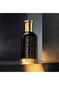 Bouteille en verre noir avec bouchon doré portant l'étiquette "BOSS Bottled Absolu", posée sur une surface gris foncé, éclairée par une lumière chaude mettant en valeur la base.
