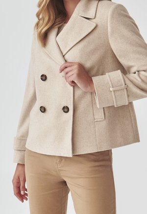 Woman wearing a beige double-breasted wool jacket with four buttons and cuff straps, paired with light brown pants.
