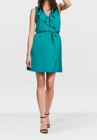 Teal sleeveless dress with ruffled neckline and cinched waist. Smooth texture, above-knee length, paired with black strappy sandals.