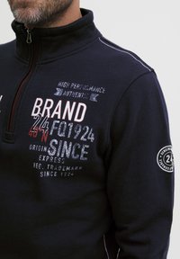 Pullover in felpa navy con colletto alto, chiusura con zip e cuciture bianche. Presenta testo stampato in rosso e bianco, oltre a una toppa rotonda sulla manica.