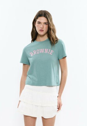 Jeune femme aux cheveux ondulés portant un t-shirt bleu sarcelle avec le texte « BROWNIE » et une jupe blanche à volants, debout devant un fond uni.