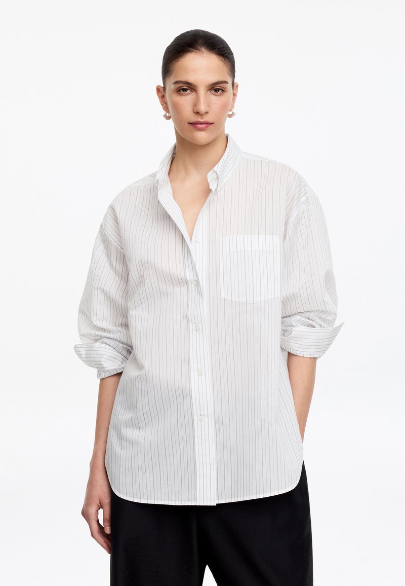 Femme aux cheveux foncés portant des boucles d'oreilles créoles dorées, une chemise blanche à fines rayures avec les manches retroussées et un pantalon noir, debout devant un fond blanc.