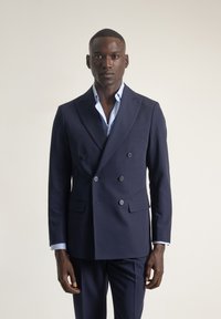 Chaqueta de traje azul marino de doble botonadura en tejido suave. Presenta solapas de pico, dos bolsillos frontales y seis botones para el cierre. Camisa de un azul claro debajo.