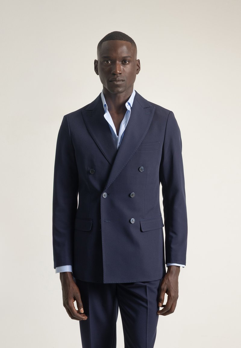 Chaqueta de traje azul marino de doble botonadura en tejido suave. Presenta solapas de pico, dos bolsillos frontales y seis botones para el cierre. Camisa de un azul claro debajo.