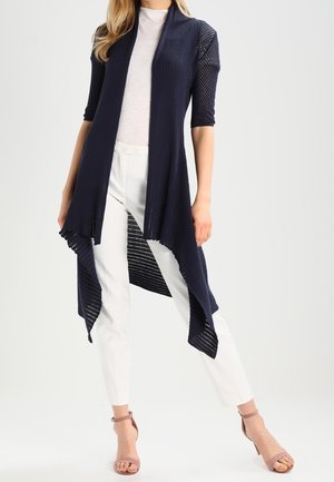 Femme portant un cardigan en maille bleu marine ouvert devant avec manches courtes, pantalon blanc taille haute et talons beige à bride à la cheville.
