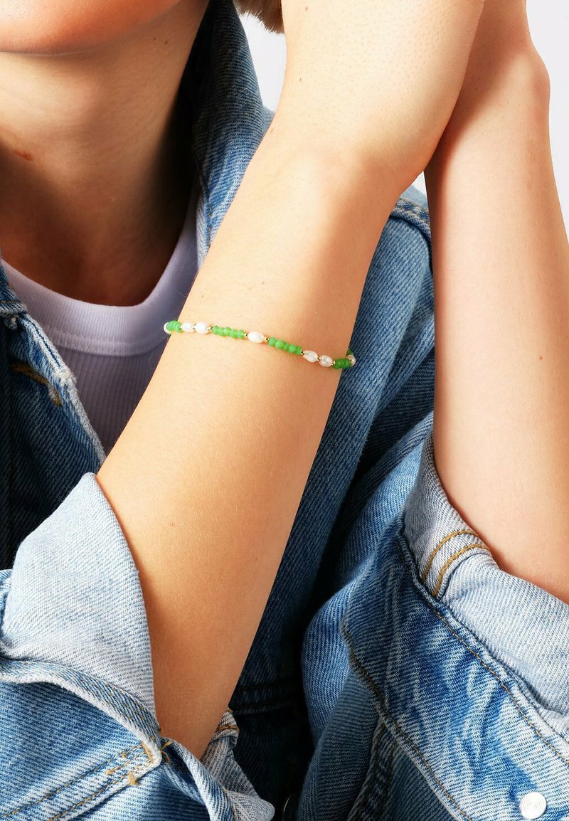 Bracciale di perline composto da perline verdi e bianche, infilato su un cordino flessibile. Indossato su un polso davanti a uno sfondo di giacca in denim.