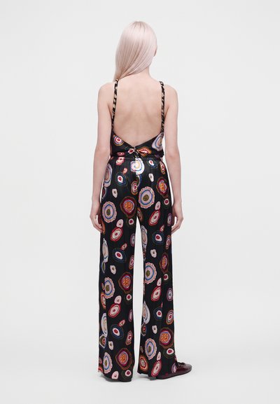 Personne aux longs cheveux clairs porte un pantalon noir à jambes larges et un haut dos nu avec des motifs circulaires colorés, debout, dos à un fond uni.