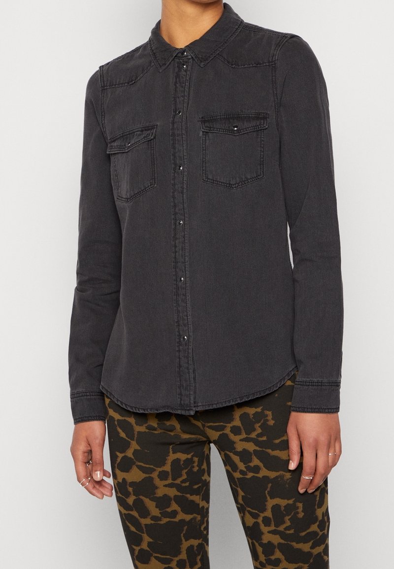 Personne portant une chemise noire à manches longues boutonnée avec deux poches poitrine et un pantalon marron et noir à motif abstrait.