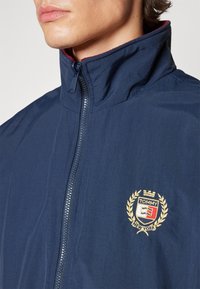 Giacca blu navy con zip, realizzata in tessuto liscio, caratterizzata da un colletto alto e un logo ricamato con accenti dorati sul petto.