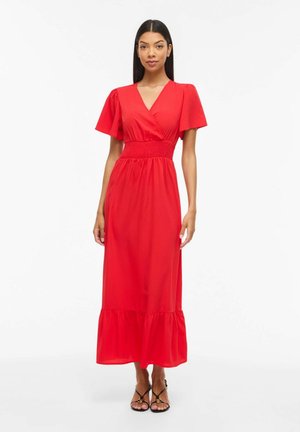 Kvinde iført en rød midi-kjole med korte flagrende ærmer, V-udskæring, taljefastholdelse og flæsekant, stående med sorte sandaler med stropper.