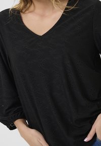 Haut noir à col en V avec un tissu texturé comportant des détails en œillets, manches 3/4 et poignets élastiqués. Matière douce et légère.