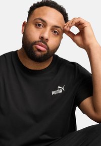Tricou din bumbac negru cu un logo alb Puma în zona stângă a pieptului, guler rotund, mâneci scurte și o textură netedă.
