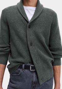 Struktur cardigan i mørkegrøn med sjalskrave, der har en knaplukning og rullede ærmer. I kombination med mørk denim jeans og en hvid skjorte.