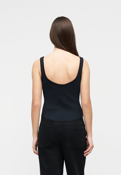 Γυναίκα με μακριά καστανά μαλλιά που φοράει μαύρο ριμπ φανελάκι με ανοιχτή πλάτη και μαύρο παντελόνι, στέκεται μπροστά σε απλό ανοιχτό φόντο.