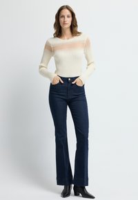 Kremowy sweter z długim rękawem w prążki z przezroczystymi akcentami, połączony z ciemnoniebieskimi dżinsami z szerokimi nogawkami w wzory. Całość dopełniają czarne botki za kostkę.