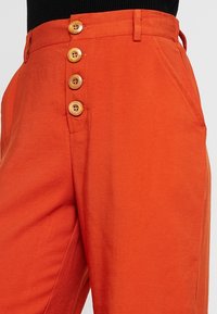 Oranje katoenen broeken met een hoge taille, zijzakken en een rij van vijf houten knopen aan de voorkant. Soepele textuur en een getailleerde pasvorm.