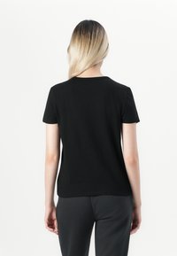 Personne avec de longs cheveux blonds portant un t-shirt noir uni à manches courtes et un pantalon sombre, se tenant de dos contre un fond blanc.