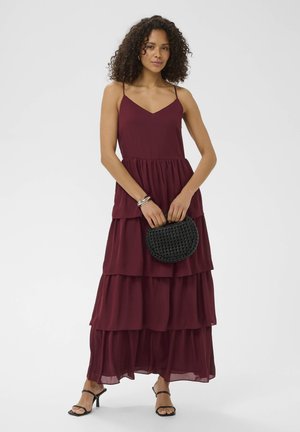 Woman wearing a sleeveless burgundy tiered maxi dress holding a black woven purse, with black strappy heels and silver bracelets.