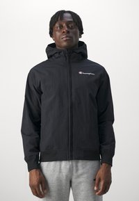 Felpa leggera nera con zip frontale e cappuccio regolabile. Presenta un piccolo logo Champion rosso e bianco sul lato sinistro del petto.