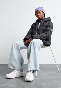 Giubbotto a vento nero, jeans a gamba larga blu chiaro e sneaker bianche. Berretto di maglia viola. Seduto su una sedia bianca contro uno sfondo chiaro.