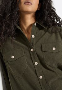 Olivgrünes Cordhemd mit langen Ärmeln, zwei Brusttaschen und hellen Knöpfen. Strukturiertes Gewebe mit einem dezenten Rippenmuster.