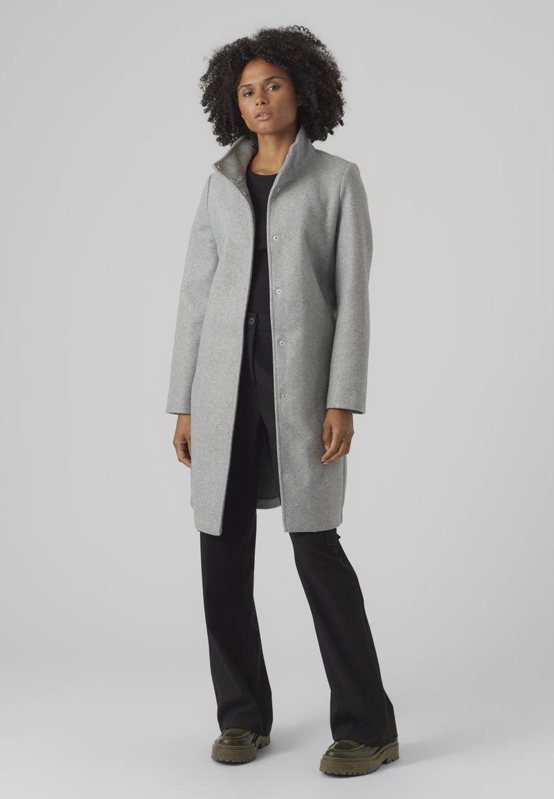 Manteau en laine gris clair avec un col haut, fermeture à boutons, et une coupe légèrement oversize, associé à un pantalon noir et des chaussures vertes.