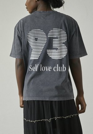 Grå oversized t-shirt med hvide stribede tal "93" og teksten "Self love club" i fed skrift på ryggen. Blødt bomuldsstof.