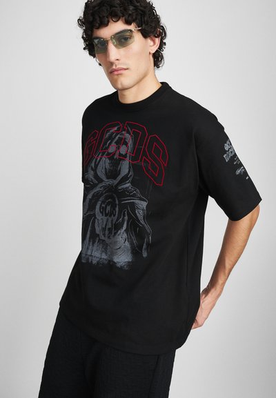 T-shirt en coton noir à manches courtes, avec un design graphique représentant une figure vêtue d'une cape et le texte "GCDS" en rouge et gris. Modèle portant des lunettes de soleil.