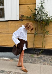 Camicia bianca a maniche lunghe, avvolto in maglia marrone, borsa a tracolla scura intrecciata e slip-on beige chiaro. In piedi su una superficie di ciottoli vicino a della vegetazione.