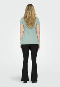 Gaiši zaļš peplum tops ar īsām piedurknēm, izplestu apakšmalu, kombinēts ar melnām platu pārslu biksēm, melna apavi, gari viļņaini mati, neitrāla fona.