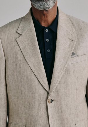 Mann mit grauem Bart, trägt ein beiges Sakko über einem schwarzen Hemd mit Knöpfen, mit einem Einstecktuch, das in der Brusttasche des Sakkos sichtbar ist.