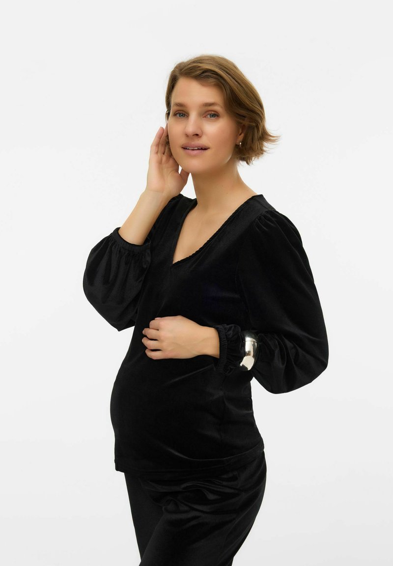 Femme enceinte en haut à manches longues en velours noir et pantalon, touchant doucement son ventre, debout devant un fond blanc uni.