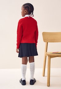 Rdeč pulover iz grobega pletenja, črna plisirana krila, bele vzorčaste nogavice do kolen in sijoči črni čevlji. Blizu preprosta lesena stol.