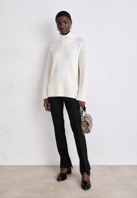Fetthaltiger weißer Rollkragenpullover mit geripptem Saum, kombiniert mit schwarzen, taillierten Hosen und einer kleinen Handtasche im Reptilienmuster. Schwarze Schuhe mit Absatz.